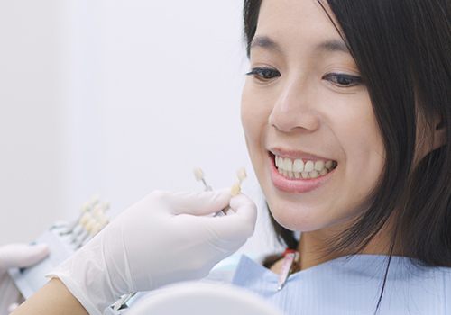 A young woman with long, dark hair smiling while a dentist uses a shade guide to determine the correct color of her metal-free dental restoration